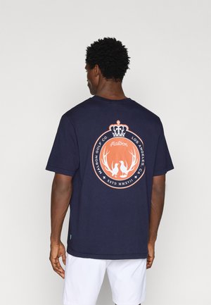 Tums zila kokvilnas t-krekls ar apaļu grafisko dizainu oranžā un baltā krāsā aizmugurē, kas attēlo ragu un krona motīvu.