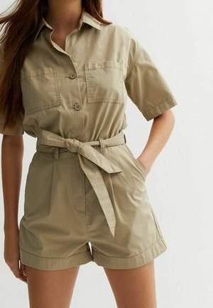 Femme portant une combinaison beige à manches courtes avec boutons, poches poitrine et ceinture nouée à la taille, debout avec une main dans la poche.