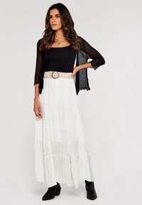 Cárdigan de encaje negro sobre una camiseta de tirantes negra ajustada, combinado con una falda maxi blanca fluida y un cinturón trenzado beige. Botines negros completan el look.