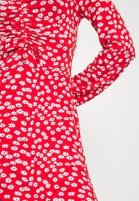 Czerwona sukienka z długim rękawem, ozdobiona wzorem białych stokrotek, z marszczonymi detalami przy biuście i dopasowanym krojem.