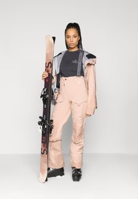 Hiihtoasu vaaleanpunaisena, jossa on säädettävät hihnat, harmaa takki, musta paita alla, pitelee suksia. Ominaisuuksiin kuuluu taskuja ja suksisiteet.