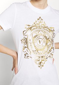 Biała bawełniana koszulka z dużym, błyszczącym złotym logo z kwiatowymi akcentami z napisem "Versace Jeans Couture" na środku.