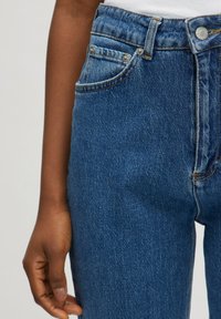 Vaqueros de mezclilla azules de cintura alta con un acabado texturizado, que cuentan con dos bolsillos frontales, acentos de botones metálicos y detalles de costura en contraste.