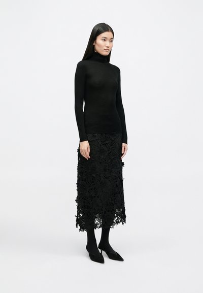 Pulover negru cu guler înalt și textură ribbed, asortat cu o fustă neagră din dantelă cu decupaje florale. Modelul poartă ciorapi negri și balerini cu vârful ascuțit.