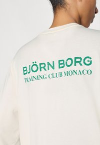 Cream short-sleeve t-shirt featuring green text that reads “BJÖRN BORG TRAINING CLUB MONACO,” with a soft texture and relaxed fit.