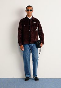 Pruun suede jakk kahe rinnakotiga ja valge kaarjas triibuga detail. Koos siniste teksade ja mustade spordijalatsitega. Hoides väikest musta kotti.