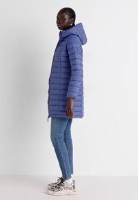 Gevoerde blauwe jas met een capuchon, voorzien van horizontale stiklijnen, lange mouwen en een rits; gecombineerd met slim-fit jeans en sneakers.