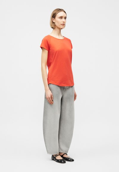 Γυναίκα που στέκεται φορώντας πορτοκαλί μπλούζα με κοντά μανίκια, ανοιχτό γκρι παντελόνι φαρδύ, και μαύρα μπαλαρίνες με ασημένιες τρύπες σε λευκό φόντο.
