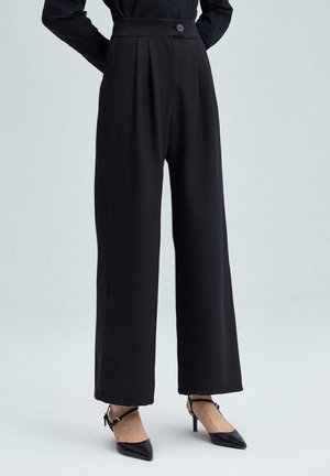 Black wide-leg trousers with a high waist, pleat detailing, a front button closure, and smooth fabric texture. Paired with black heeled shoes.