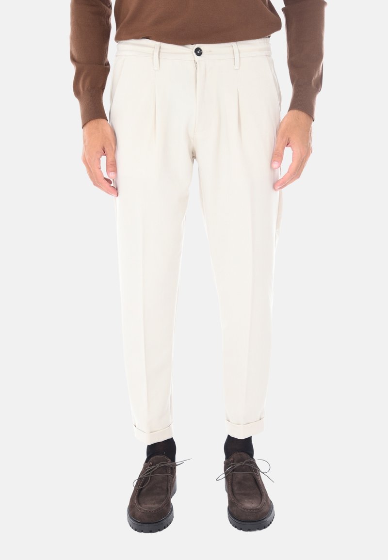 Pantaloni beige a pieghe con vestibilità rilassata, dotati di chiusura a bottone nera, orli risvoltati, indossati con scarpe marroni scure e calzini neri.