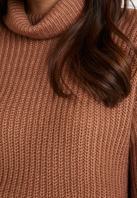 Pull à col roulé brun en tricot avec une texture côtelée. Présente un large col rabattu et une coupe décontractée.