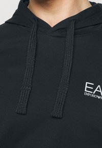 Detailní záběr na černou mikinu s kapucí s hustými copánkovými šňůrkami a bílým logem Emporio Armani EA na hrudi.