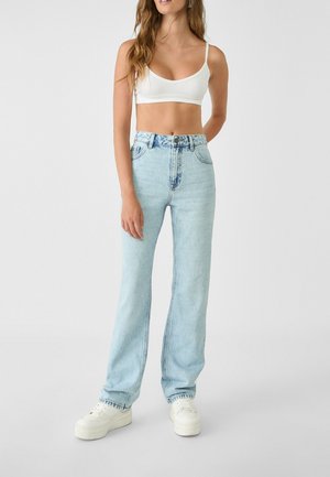Femme portant un crop top blanc, un jean taille haute bleu clair et des baskets à plateforme blanches, debout devant un fond uni.