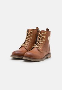 Botas de tobillo de cuero marrón con ojales dorados, cordones beige, cremallera lateral y suela de goma texturizada; el diseño presenta un acento cosido en el lateral.