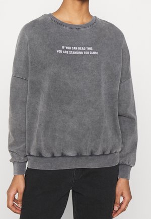 Personne portant un sweat-shirt gris délavé avec un texte blanc sur la poitrine indiquant "IF YOU CAN READ THIS YOU ARE STANDING TOO CLOSE."