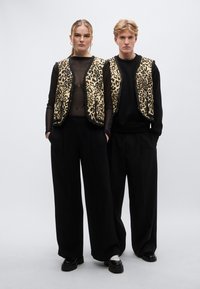 Két modell fekete ruhákat visel, bézs alapon leopárdmintás mellénnyel. A mellényeken lekerekített nyakkivágás és gombok nélküli kialakítás látható, mely kiemeli a merész mintákat.