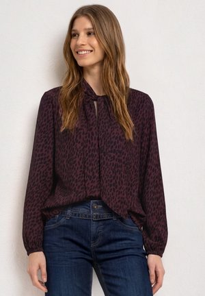 Femme aux longs cheveux châtain clair, souriante, portant un chemisier violet foncé à taches noires et un jean bleu sur un fond uni.