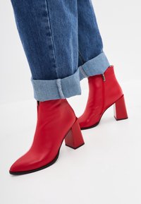 Bottines en cuir rouge avec un bout pointu et un talon carré. Elles disposent d'une fermeture éclair latérale et d'une texture lisse et brillante. Le jean est retroussé à la cheville.