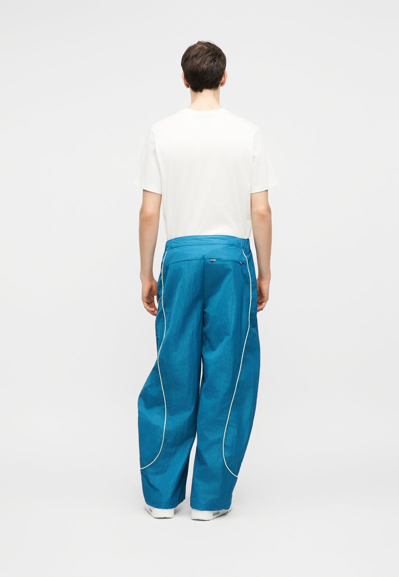 Uomo che indossa pantaloni larghi blu con piping bianco e una maglietta bianca a maniche corte, in piedi con la schiena rivolta verso l'avanti su sfondo bianco.