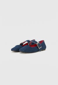 Ballerines en denim bleu avec un bout pointu, un détail de sangle effiloché et une doublure intérieure rouge ; elles possèdent une petite boucle en métal et une semelle plate.