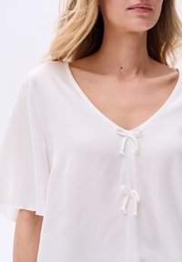 Biała, lekka bluzka z dekoltem w serek, krótkimi rękawami i dekoracyjnymi kokardkami przy dekolcie. Gładka tekstura, minimalne detale szwów.