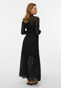 Vestido largo negro de encaje, con mangas largas y cuello alto. El diseño presenta una textura transparente con una silueta fluida.