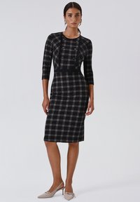 Zwart-crèmekleurige geruite jurk met driekwart mouwen, een aangesloten silhouet, knopen aan de voorkant en een zoom tot op de knie. Paar beige pumps met hak.