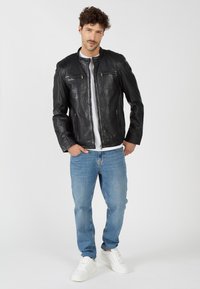 Schwarze Lederjacke mit Stehkragen und Reißverschluss, versehen mit zwei Brusttaschen und einer glatten Textur, getragen mit blauen Jeans und weißen Sneakers.
