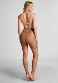 Frau in einem hautfarbenen BH, hochgeschnittenen Formshorts und hautfarbenen High Heels, die mit dem Rücken zur Kamera auf einem grauen Hintergrund posiert.