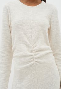 Långärmad krämfärgad topp i texturerad tyg, med en central rynkdetalj och rund ringning, vilket skapar en figurnära silhuett.