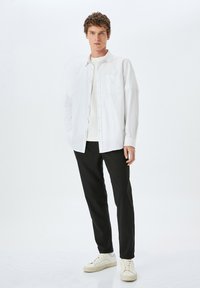 Witte button-up shirt over een lichte trui, gecombineerd met zwarte broek en witte sneakers. Het shirt heeft een zak en lange mouwen.