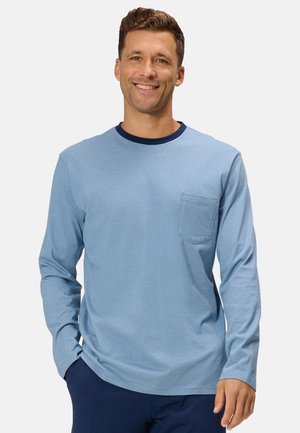 Smiling man wearing a light blue long-sleeve shirt with a chest pocket and dark blue pants, standing with one hand in his pocket.