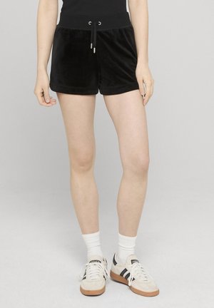 Persoon in zwarte fluwelen korte broek met trekkoord, witte sportsokken en witte Adidas sneakers met zwarte strepen, staand op een grijze vloer.