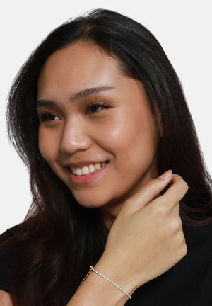 Gouden armband met een rij heldere rhinestones. Vrouw met lang donker haar draagt een zwarte top en glimlacht terwijl ze de armband aanraakt.