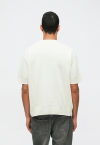 Mann som vender bort fra kameraet iført en hvit short-sleeve genser og falmede sorte jeans mot en lys, ensfarget bakgrunn.