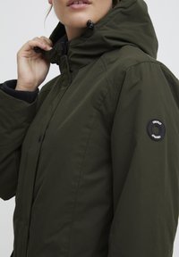 Parka olive verte avec fermeture éclair et bouton, capuche ajustable et un patch rond sur la manche. Tissu doux et texturé.