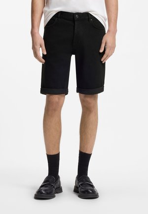 Mand iført sorte shorts med optrukket kant, sorte sokker og sorte lædersko, stående mod en ensfarvet hvid baggrund.