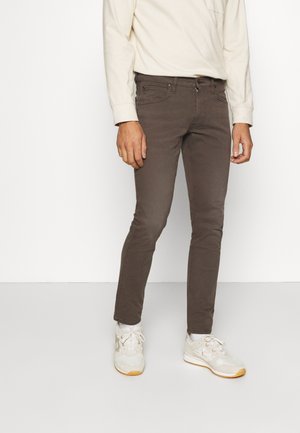 Mann trägt braune, eng anliegende Hose, beiges langärmeliges Hemd und weiße Turnschuhe, steht vor weißem Hintergrund.