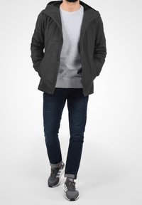 Harmaa kevyt takki hupulla, yhdistettynä harmaaseen neuleeseen, tummansinisiin farkkuihin ja harmaisiin urheilukengiin, seisten.