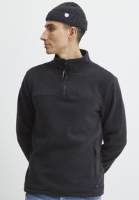 Temno siva flis majica s četrtinskim zadrgo, visokim ovratnikom, dvema stranskima žepoma in malo nalepko z logotipom na levi strani.