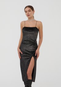 Abito midi nero glitterato con sottili spalline a spaghetti, caratterizzato da un design arricciato sul lato e uno spacco alta coscia per un dettaglio aggiuntivo.