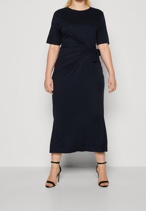 Femme portant une robe midi noire à manches courtes avec un détail à nouer sur le côté, associée à des sandales à talons hauts noires avec bride à la cheville.