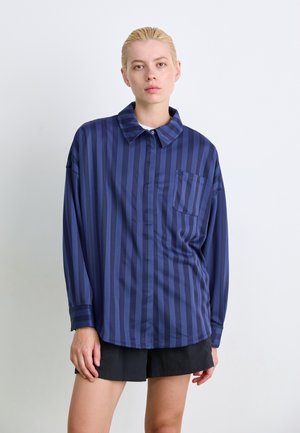 Navy blue long-sleeve shirt with vertical stripes, soft fabric, button collar, front pocket, and a relaxed fit. Paired with black shorts.