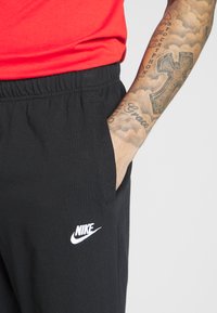 Črne trenirke z elastičnim pasom, stranskimi žepi in belim logotipom Nike. Gladka tekstura blaga. Rdeča majica vidna nad pasom.