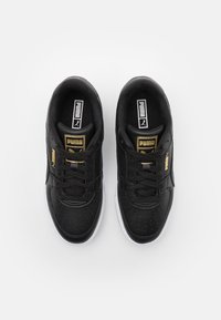 Zapatillas de cuero negro con textura perforada, acentos dorados en el lateral, suelas de goma blanca y cordones negros. Logo de Puma en la lengüeta.