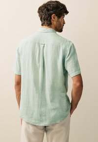 Short-sleeve button-up shirt in light green textured fabric, featuring a back pleat and rounded hem, standard collar, and rolled cuffs.