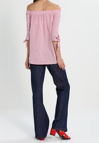 Donna che indossa una blusa a strisce rosa con spalle scoperte e maniche annodate, jeans a gamba larga scuri e sandali con tacco blocco aperti rossi, in piedi con la schiena visibile.