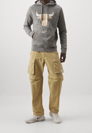 Man wearing gray hoodie with bull logo, beige cargo pants, and white sneakers with black Nike swoosh, standing against plain background.