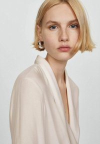 Top avvolgente di un beige chiaro con scollatura a V profonda, tessuto morbido e maniche a tre quarti. Modella che indossa grandi orecchini a cerchio in argento.