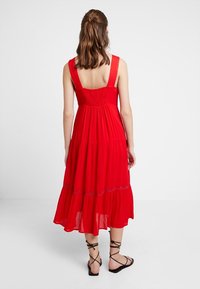 Robe maxi rouge à volants avec un corsage smocké, de larges bretelles et des accents décoratifs bleus. Tissu léger avec une coupe fluide.